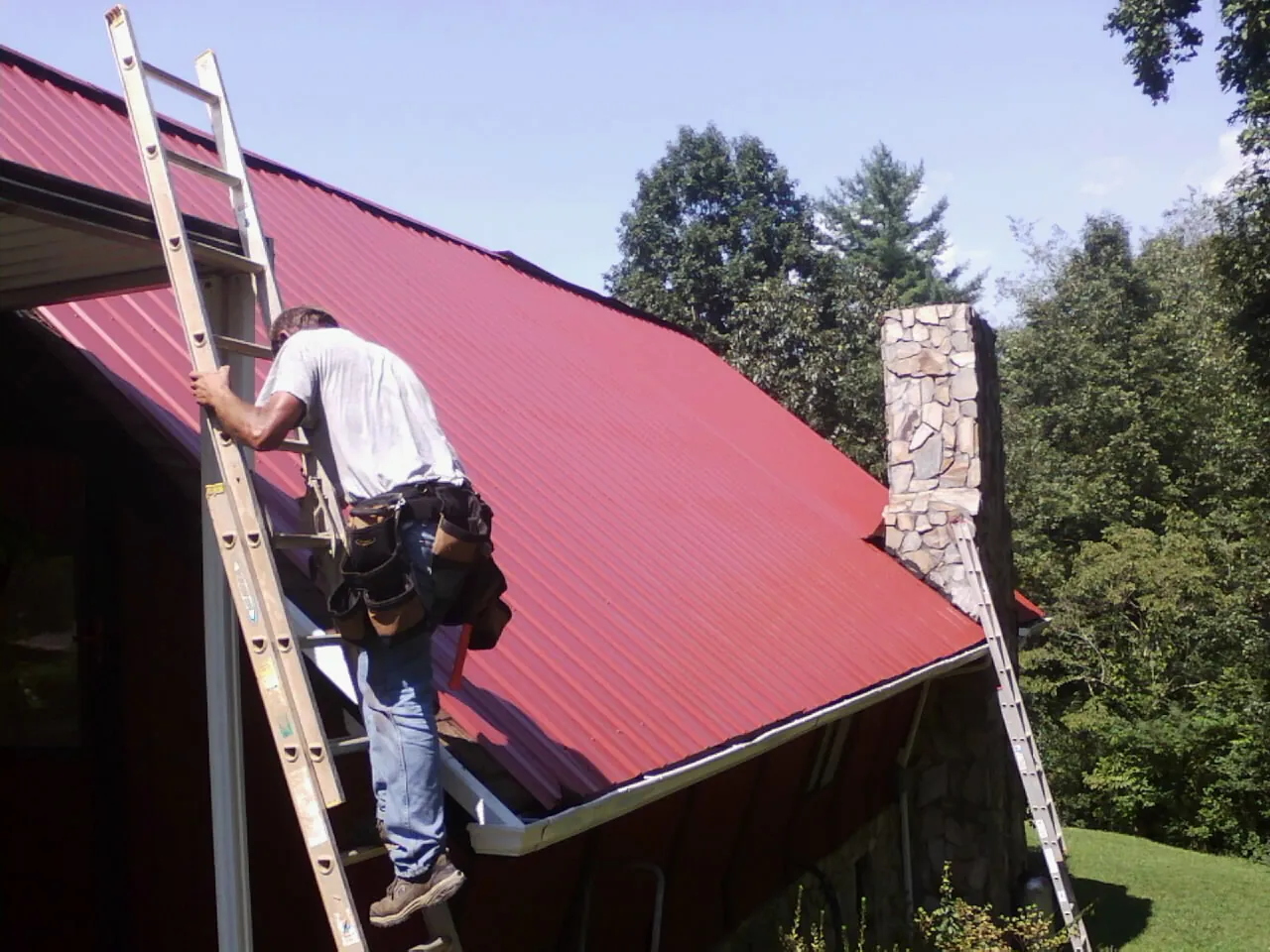 Roofer on a ladder beside a red metal roof and stone chimney — Soffit & Fascia Repair by Roofing Brothers in Briarcliff Manor